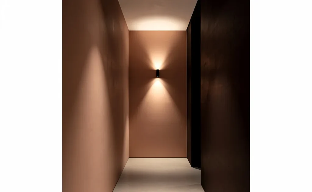 Un couloir étroit et peu éclairé présente un mur en terracotta clair et l’autre en terracotta foncé, avec une applique murale projetant des ombres douces et une ambiance chaleureuse.