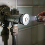 Un mécanisme ancien de chasse d'eau Porcher est éclairé par une main tenant une petite lampe torche dans une salle de bains carrelée d'époque.